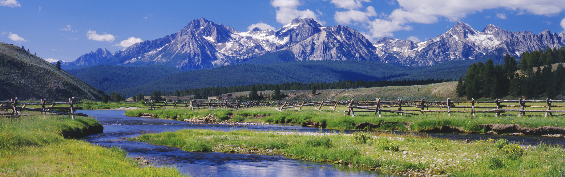 A serene landscape with a clear stream, green grass, and a wooden fence. Snow-capped mountains are in the distance.