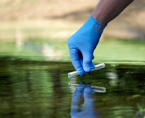 A gloved hand lowers a test tube into lake water.